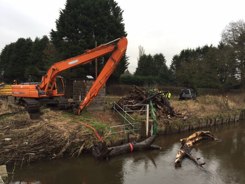 large digger on river bank clearing trees which have fallen